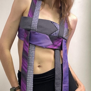 以下が含まれることがあります： 紫、黒、グレーの色合いのネクタイを複数組み合わせて作られたクロップトップ。ストライプ柄やドット柄が混ざり合っており、身頃やショルダーストラップからネクタイの端が数本垂れ下がっているデザインの衣類。