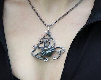 May include: A silver octopus pendant necklace. The pendant features an octopus design with swirling tentacles and a small, round gemstone. The necklace has a dark chain and is worn against a black top.