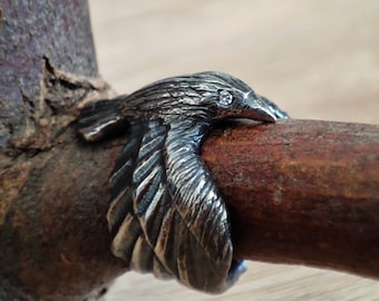 May include: A silver ring with a raven design, featuring a small gemstone eye. The raven is perched on a wooden branch. The ring's design includes detailed feathers and the bird's head, creating a striking piece of jewellery.