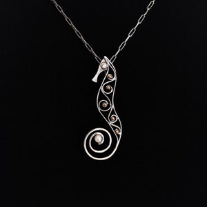 Puede incluir: Un collar con colgante de caballito de mar plateado. El dise&ntilde;o del caballito de mar presenta una cola en espiral y est&aacute; adornado con peque&ntilde;os acentos redondos de color dorado y una perla. El collar cuelga de una cadena plateada sobre un fondo negro.
