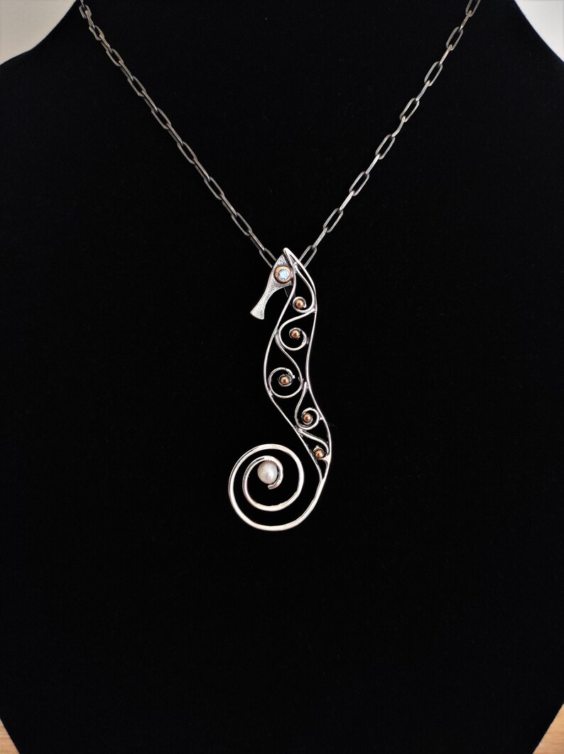Puede incluir: Un collar con colgante de caballito de mar plateado. El dise&ntilde;o del caballito de mar presenta una cola en espiral y est&aacute; adornado con peque&ntilde;os acentos redondos de color dorado y una perla. El collar cuelga de una cadena plateada sobre un fondo negro.