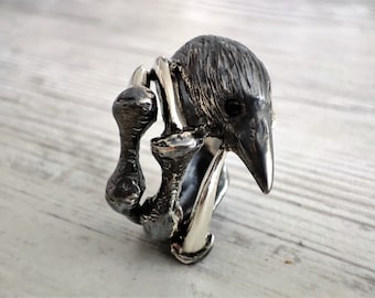 May include: A silver-toned ring featuring a stylized bird design. The ring has a textured finish, with the bird's head and beak prominently displayed. The ring's design includes a curved, open band and a detailed, three-dimensional bird.