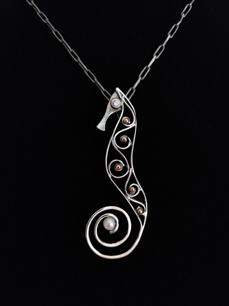 Puede incluir: Un collar con colgante de plata con forma de caballito de mar con un dise&ntilde;o calado delicado. El caballito de mar presenta una peque&ntilde;a gema redonda de color azul claro y varias peque&ntilde;as cuentas de color cobre. Una perla est&aacute; en la base. La cadena es de plata.