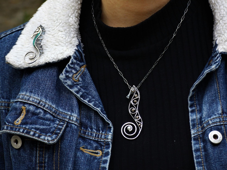 Puede incluir: Un collar con colgante de caballito de mar plateado y un broche a juego. El colgante de caballito de mar presenta una cola en espiral y est&aacute; adornado con peque&ntilde;as cuentas plateadas. El broche tiene un acento turquesa. La joyer&iacute;a se muestra sobre una camiseta negra y una chaqueta vaquera.