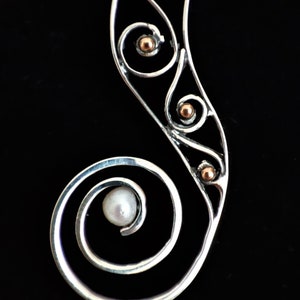 Puede incluir: Colgante plateado con dise&ntilde;o en espiral, con una perla y peque&ntilde;os detalles dorados. El colgante tiene una forma curva y alargada, sobre un fondo negro.
