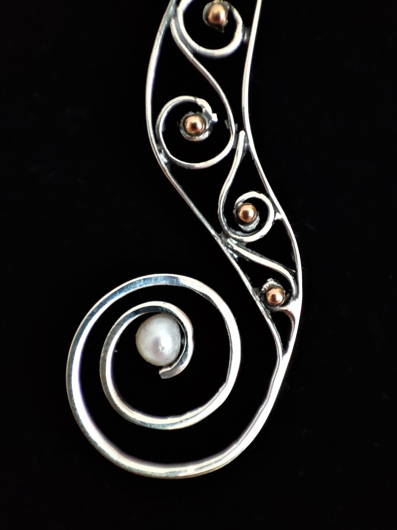 Puede incluir: Colgante plateado con dise&ntilde;o en espiral, con una perla y peque&ntilde;os detalles dorados. El colgante tiene una forma curva y alargada, sobre un fondo negro.