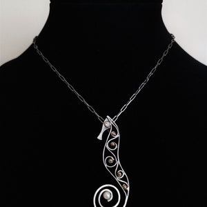 Puede incluir: Un collar con colgante de plata con forma de caballito de mar. El dise&ntilde;o del caballito de mar presenta una cola en espiral y est&aacute; adornado con peque&ntilde;os acentos redondos de color dorado y una perla. El collar se exhibe en un maniqu&iacute; negro.