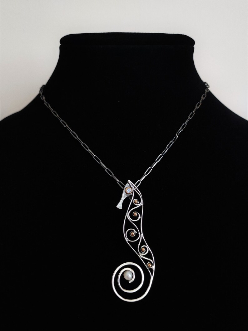 Puede incluir: Un collar con colgante de plata con forma de caballito de mar. El dise&ntilde;o del caballito de mar presenta una cola en espiral y est&aacute; adornado con peque&ntilde;os acentos redondos de color dorado y una perla. El collar se exhibe en un maniqu&iacute; negro.