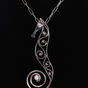 Puede incluir: Collar con colgante de plata con forma de caballito de mar. El dise&ntilde;o del caballito de mar presenta un ojo de piedra azul, detalles dorados y una perla en la cola. El colgante cuelga sobre un fondo negro.