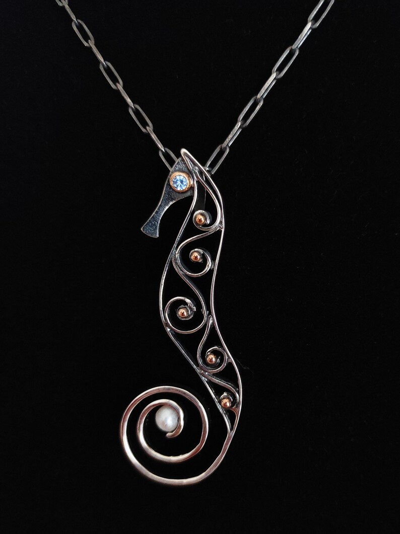 Puede incluir: Collar con colgante de plata con forma de caballito de mar. El dise&ntilde;o del caballito de mar presenta un ojo de piedra azul, detalles dorados y una perla en la cola. El colgante cuelga sobre un fondo negro.
