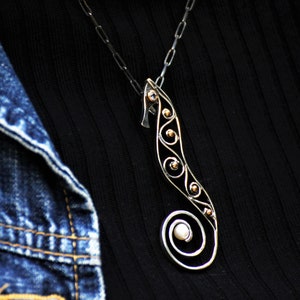 Puede incluir: Un collar con colgante de plata con forma de caballito de mar de dise&ntilde;o delicado. El caballito de mar presenta una cola en espiral y est&aacute; adornado con peque&ntilde;os detalles dorados y una perla. El collar se lleva sobre una camiseta negra y una chaqueta vaquera.
