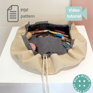 Pode incluir: Uma bolsa de maquiagem bege com cordão e forro listrado cinza e branco. A bolsa está aberta e cheia de vários itens de maquiagem, incluindo pincéis, batons e paletas de sombras para os olhos. A bolsa está sobre uma superfície branca com um logotipo branco e azul que diz "Ella Sewing Studio" e uma superfície rosa no fundo. O texto "PDF pattern" e "Video tutorial" também é visível na imagem.