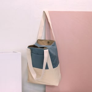 May include: A blue and cream canvas tote bag with a single strap and a brown fabric lining. The bag is hanging on a pink wall.