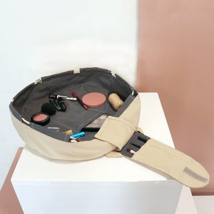 Puede incluir: Una bolsa de maquillaje beige y gris con cierre de cremallera y una bolsa separada para pinceles. La bolsa está abierta y llena de artículos de maquillaje, incluyendo colorete, lápiz labial, máscara de pestañas y base de maquillaje.