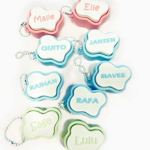 Peut inclure: Plusieurs porte-clés en forme de nuage aux couleurs pastel, chacun avec un nom différent dans une police assortie. Les porte-clés sont roses, bleus et verts, avec des noms comme Malie, Elle, Quito et Jansen. Chaque porte-clés a une chaîne argentée.