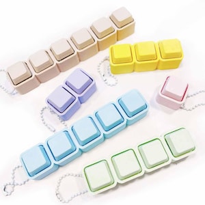 May include: A collection of colorful, square-shaped pill organizers in various pastel shades, including beige, yellow, lavender, light blue, and pink. Each organizer is connected by a chain, and the design resembles keyboard keys. The background is white.