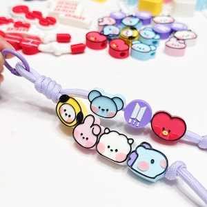 May include: A light purple lanyard with cartoon character charms in various colors. The charms include a yellow bear, pink bunny, white bear, blue bear, purple logo, and red heart. Other charms are scattered in the background.