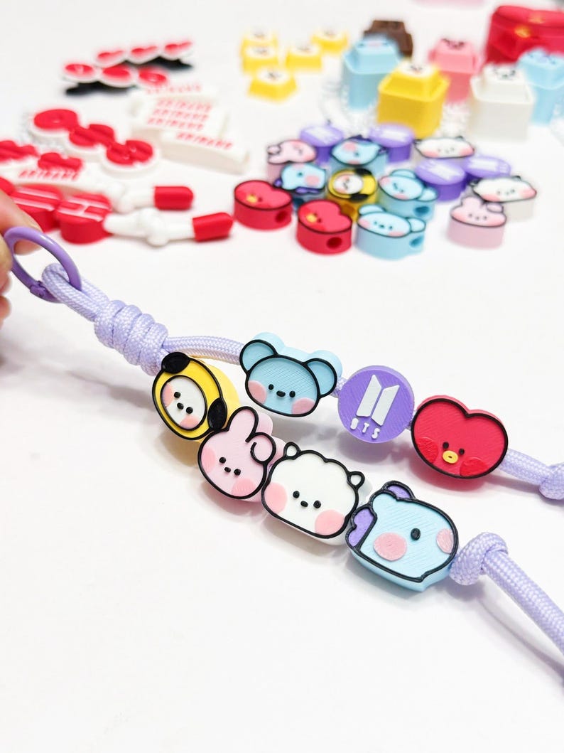 May include: A light purple lanyard with cartoon character charms in various colors. The charms include a yellow bear, pink bunny, white bear, blue bear, purple logo, and red heart. Other charms are scattered in the background.