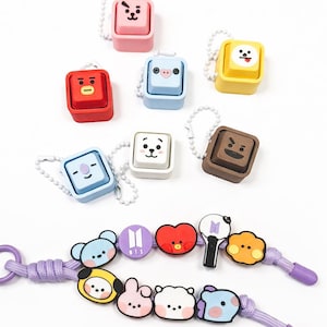 May include: A selection of colourful character keychains and a purple lanyard with cartoon figures. The keychains are square-shaped with various facial expressions. The lanyard features multiple cartoon characters and the BTS logo.