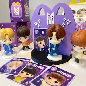 May include: A collection of Tiny Tan figurines and cards. The figurines have various hair colours and outfits. The cards feature character illustrations with names like J-Hope, Suga, and Jimin. Purple McDonald's-themed boxes and a display stand are also visible.