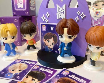 May include: A collection of Tiny Tan figurines and cards. The figurines have various hair colors and outfits. The cards feature character illustrations with names like J-Hope, Suga, and Jimin. Purple McDonald's-themed boxes and a display stand are also visible.