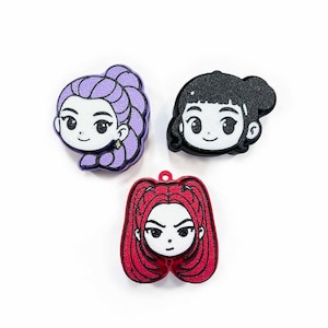 May include: Three cartoon character charms with different hairstyles and colors. One has purple hair, another has black hair, and the third has red hair. Each charm has a white face with black facial features and a small loop at the top.