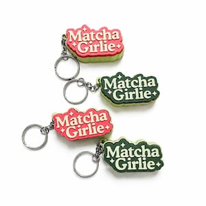 Puede incluir: Cuatro llaveros con un diseño suave y elevado en forma de nube con el texto "Matcha Girlie" en una fuente serif. Dos llaveros son de color rosa con texto crema y dos son de color verde oscuro con texto crema, decorados con iconos de estrellas.