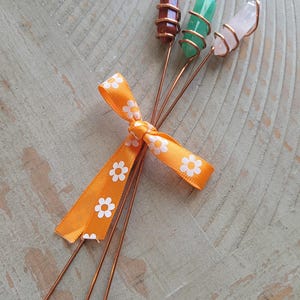 May include: Three crystal skewers with copper wire wrapping, featuring brown, green, and clear stones. An orange ribbon with white daisy patterns is tied around the skewers. The skewers are arranged on a light-colored wooden surface.