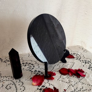 May include: A black circular mirror with a decorative pattern sits on a black stand. The mirror is surrounded by red rose petals on a white lace tablecloth.