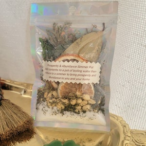 May include: A clear, iridescent pouch filled with dried herbs, spices, and citrus slices. The pouch contains a bay leaf, dried orange slices, and other ingredients. Text on the label reads "Prosperity & Abundance Simmer Pot".