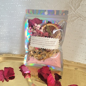 May include: A clear, iridescent pouch filled with dried flowers, herbs, and a dried orange slice. The pouch contains a pink powder and is labelled "Love Simmer Pot". Scattered rose petals are in the foreground.