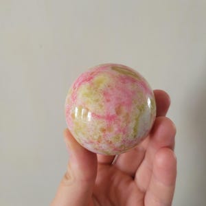 May include: A polished sphere with a mix of pink and yellow hues. The stone ball is held in a hand, showcasing its smooth, reflective surface. The sphere's colours blend together, creating a marbled effect.