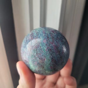 May include: A hand holds a polished sphere of ruby zoisite. The stone is a mix of deep teal, green, and magenta hues, with a speckled appearance. The sphere is smooth and reflective, showcasing the natural patterns of the stone.