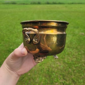Peut inclure: Un petit pot rond en laiton poli doté de deux poignées latérales texturées et ornées, ainsi que de trois pieds décoratifs. La surface métallique présente une teinte jaune doré chaleureuse avec des reflets variés et un bord supérieur lisse.