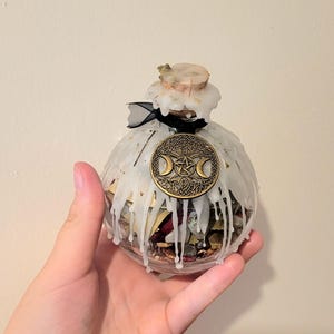 May include: A spherical glass bottle filled with dried herbs and crystals. It has a cork stopper and a gold-coloured metal emblem with a triple moon design. White wax drips down the sides, and a black ribbon is tied around the neck.