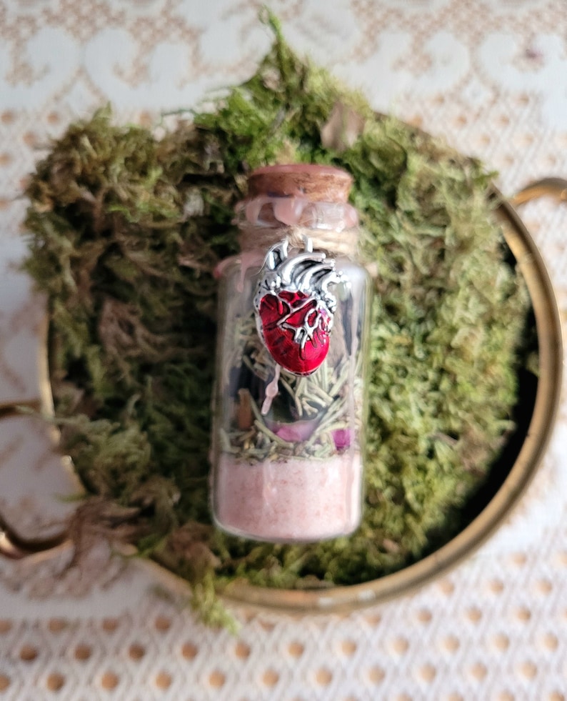 Puede incluir: Una peque&ntilde;a botella de vidrio llena de hierbas, cristales y un dije de coraz&oacute;n rojo. La botella est&aacute; coronada con un corcho y tiene un alambre plateado atado alrededor.