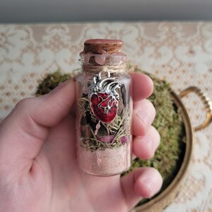 Puede incluir: Una peque&ntilde;a botella de vidrio con un tap&oacute;n de corcho llena de hierbas secas, sal y un dije de coraz&oacute;n rojo. La botella est&aacute; atada con un cordel y tiene un sello de cera.