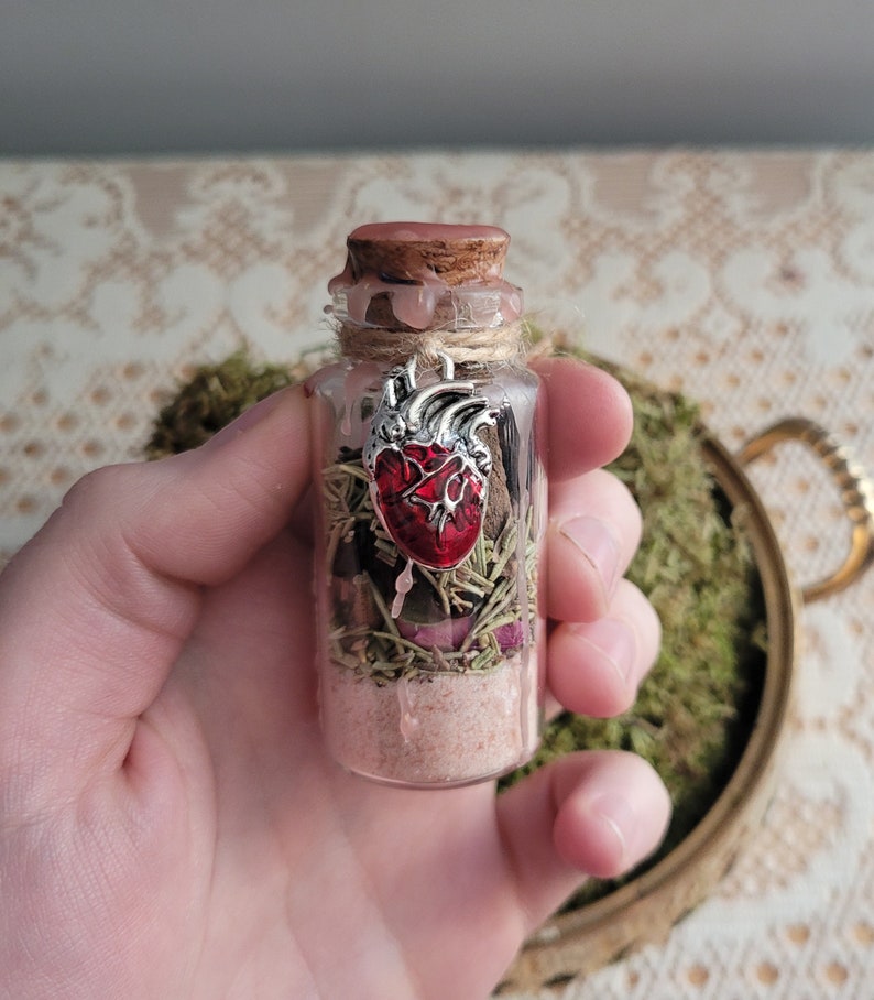 Puede incluir: Una peque&ntilde;a botella de vidrio con un tap&oacute;n de corcho llena de hierbas secas, sal y un dije de coraz&oacute;n rojo. La botella est&aacute; atada con un cordel y tiene un sello de cera.