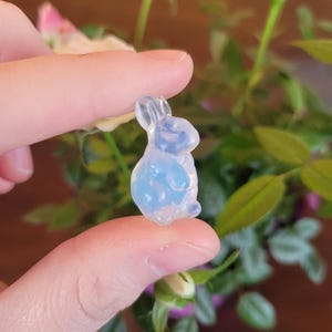 May include: A small, translucent blue and white bunny figurine. The bunny is held between fingers, with a blurred background of green leaves and pink and white rose buds. The figurine is a miniature sculpture.