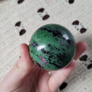 May include: A polished sphere of green zoisite with black and ruby inclusions. The gemstone ball is held in a hand, showcasing its smooth, reflective surface. The sphere's colours and patterns create a striking visual effect.