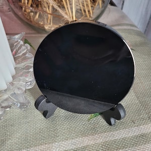 May include: A black, round object with a shiny surface, resting on a small, black stand. The stand has a three-legged design. The object appears to be a lid or a decorative piece.