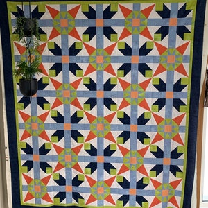 Pode incluir: Uma colcha com um padrão geométrico de estrelas em tons de azul, verde, laranja e branco. A colcha está pendurada em uma parede com uma borda azul escura.