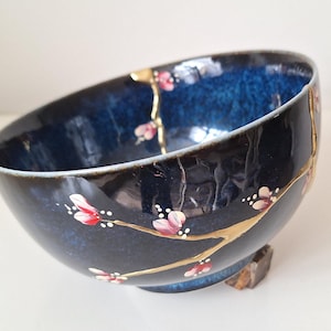 Può includere: Una ciotola in ceramica blu scuro con un motivo floreale dorato e rosa. La ciotola presenta una crepa nella smaltatura, che è evidenziata da una vernice dorata.