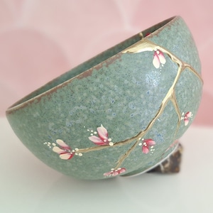 Puede incluir: Un pequeño cuenco de cerámica verde con un esmalte craquelado y detalles dorados. El cuenco está decorado con un diseño pintado a mano de una rama con flores rosas.