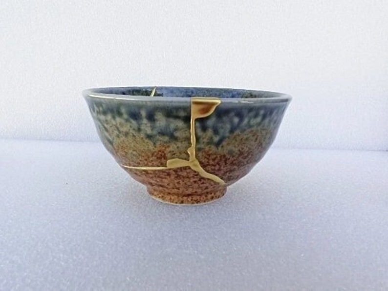 May include: A small, blue and brown ceramic bowl with a crack filled with gold paint. The bowl is slightly chipped on the rim.