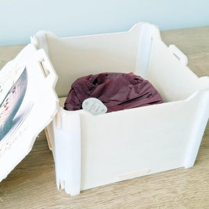 May include: A white box with a hinged lid. The lid has a floral design and the words "Happy Birthday" printed on it. The box is open and a purple fabric item is inside.