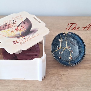 Puede incluir: Una caja blanca con una tapa que presenta un dise&ntilde;o floral y el texto "The Art of Kintsugi" y "Kanelasuuri". La tapa est&aacute; abierta y se ve un peque&ntilde;o cuenco de cer&aacute;mica azul y dorado con una grieta reparada con oro.