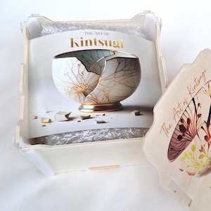 Puede incluir: Una caja de madera con tapa, que contiene un libro titulado "The Art of Kintsugi". El libro presenta un taz&oacute;n de cer&aacute;mica blanco y dorado con grietas reparadas con oro. La tapa de la caja tiene una imagen similar de un taz&oacute;n con acentos dorados.