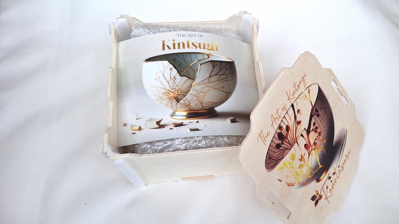 Puede incluir: Una caja de madera con tapa, que contiene un libro titulado "The Art of Kintsugi". El libro presenta un taz&oacute;n de cer&aacute;mica blanco y dorado con grietas reparadas con oro. La tapa de la caja tiene una imagen similar de un taz&oacute;n con acentos dorados.