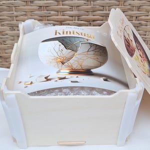 May include: A white wooden box with a lid. The lid has a printed image of a broken bowl with gold accents. The box is labeled "The Art of Kintsugi".